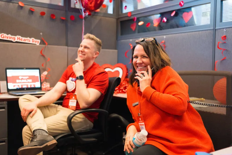Two smiling staffers in red attire at a heart-decorated office cubicle during Giving Hearts Day