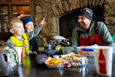 Family of three laughing over a meal inside a rustic ski lodge with a stone fireplace