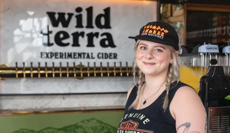 Smiling bartender in a black cap and tank top standing in front of a Wild Terra Experimental Cider tap wall