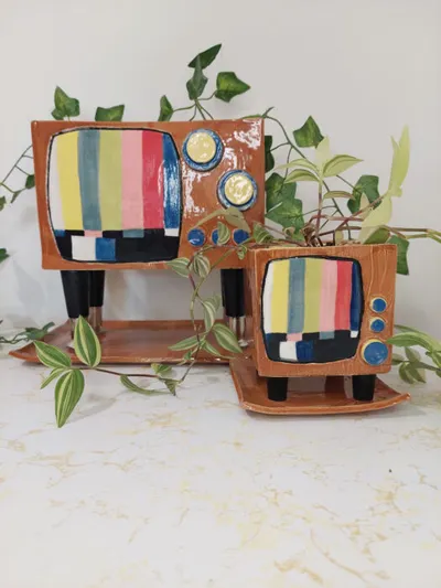 Two terracotta ceramic planters shaped like retro TVs with colorful test-pattern screens, trailing green ivy