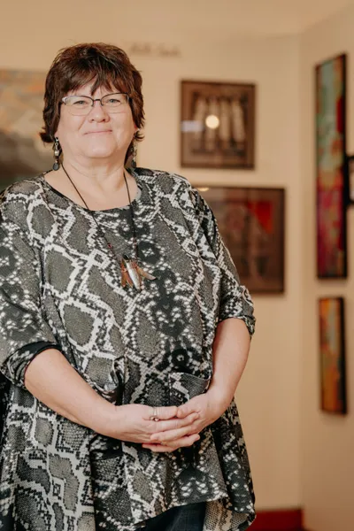Woman in snake-print top wearing a long pendant necklace stands in a gallery with framed artwork behind her