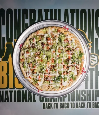 Pizza topped with lettuce, tomatoes, and white dressing on a metal pan over a championship congratulations banner