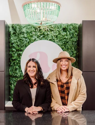 Two smiling women leaning on a counter in front of a green living wall and beaded chandelier