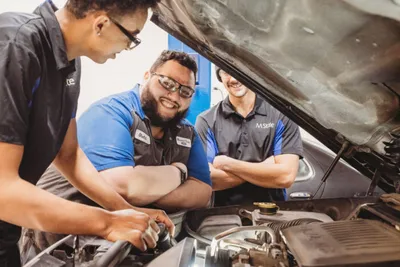 Three auto mechanics in blue uniforms examine an open car engine together