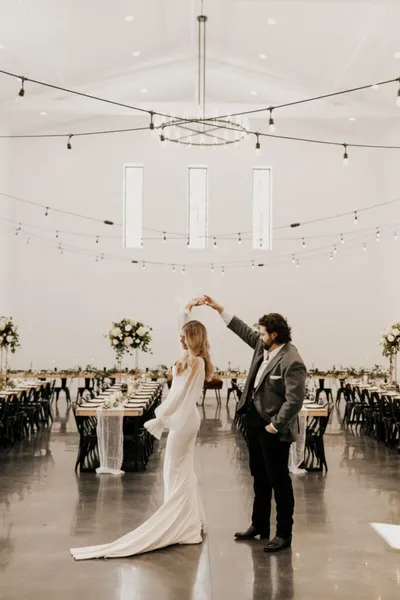 Couple dancing in a bright white reception hall with string lights and set banquet tables