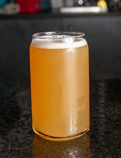 Hazy golden beer with a white foam head in a can-shaped glass on a dark bar top