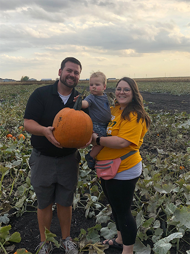 Man holding large orange pumpkin stands with woman and toddler in a pumpkin patch field