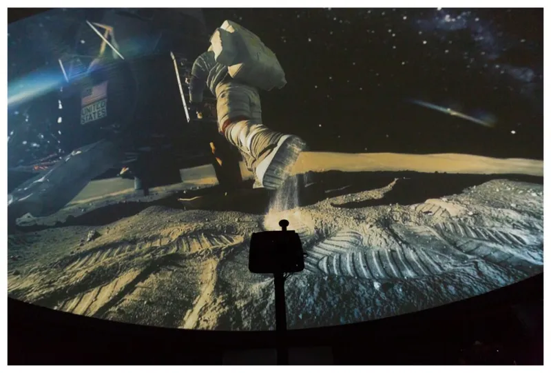 Planetarium dome screen showing an astronaut on the lunar surface with a silhouetted projector in the foreground