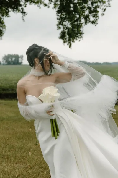 Bride in white strapless gown and flowing veil holds white bouquet in an open field