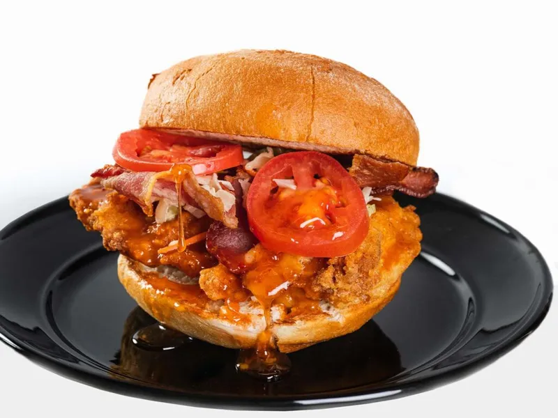 Fried chicken sandwich with bacon, tomato slices, coleslaw, and orange sauce on a black plate
