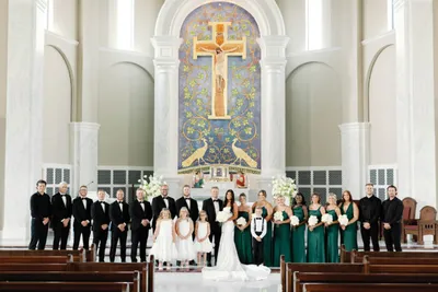 Wedding party posed at a church altar, bridesmaids in emerald green gowns, groomsmen in black tuxedos, gold cross and mosaic