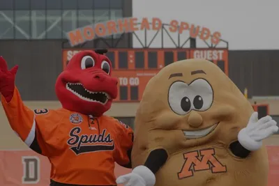 Moorhead Spuds mascot and a potato mascot with an M logo pose in front of a scoreboard