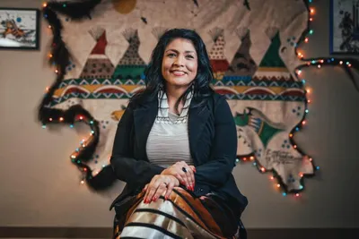 Woman in black blazer smiling beside a drum, with a colorful tipi-patterned mural and string lights behind her
