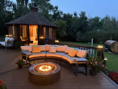 Curved outdoor sectional sofa around a glowing fire pit on a wood deck, with a lit gazebo at dusk