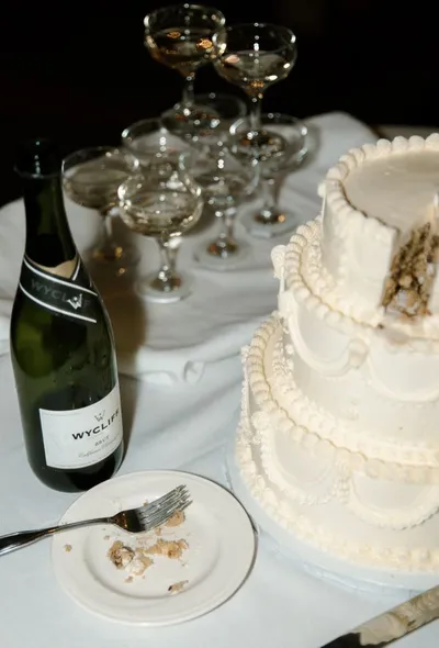 Three-tier white wedding cake beside a sparkling wine bottle and coupe glasses