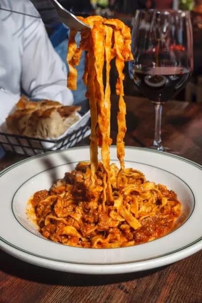 Fork lifting ribbon pasta in rich meat sauce over a white plate, red wine and bread in background