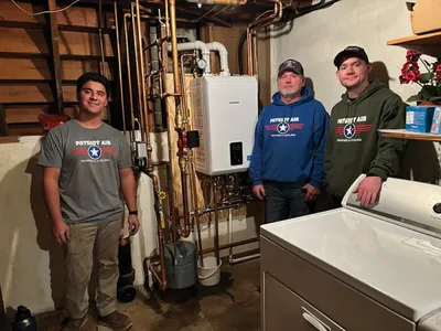 Three technicians in matching branded shirts standing beside a newly installed tankless water heater in a basement utility