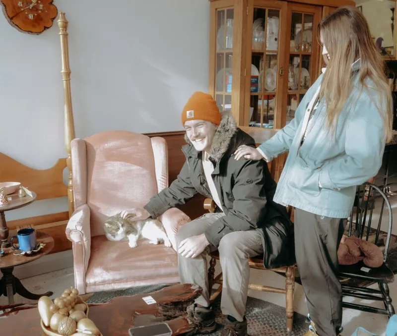 Man in orange beanie petting a cat on a pink armchair while a woman looks on in a consignment shop