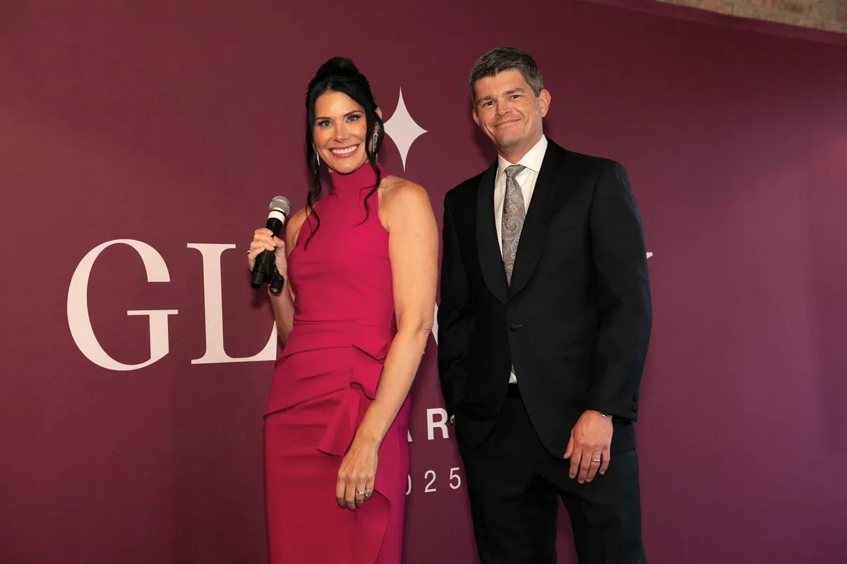 Woman in pink dress holding microphone poses with man in dark suit before a magenta branded backdrop