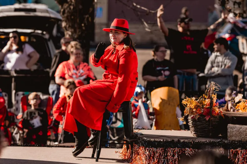 Woman in red coat and wide-brim hat waves from a fall-decorated parade float