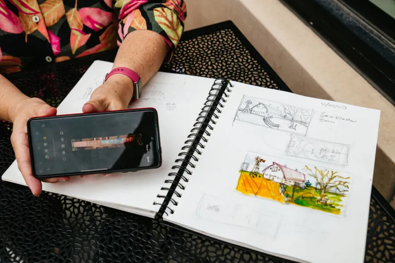 Person holding a smartphone beside an open spiral sketchbook showing pencil sketches and a watercolor farm scene
