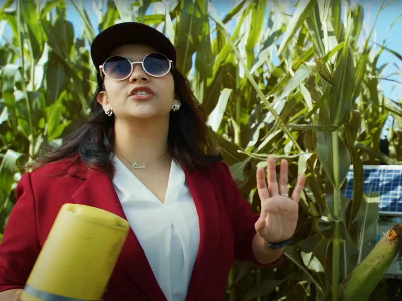 Woman in red blazer and sunglasses gestures while standing before tall corn stalks