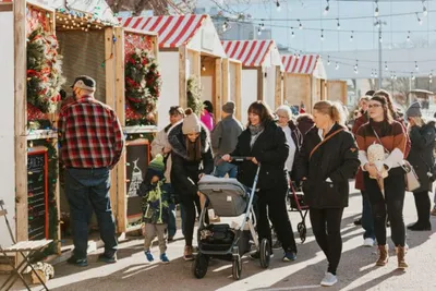 Crowd of shoppers browsing outdoor holiday market stalls with red-striped awnings and wreaths
