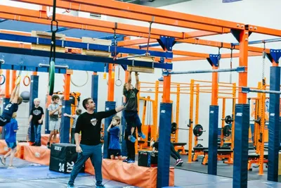 Person on monkey bars in a ninja warrior-style gym with orange and blue equipment