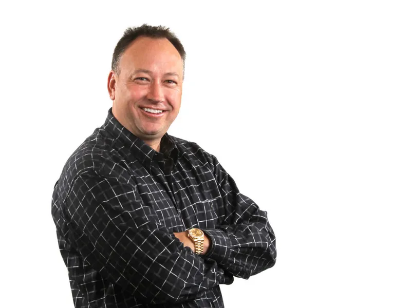 Smiling man with arms crossed wearing a dark plaid shirt and gold watch