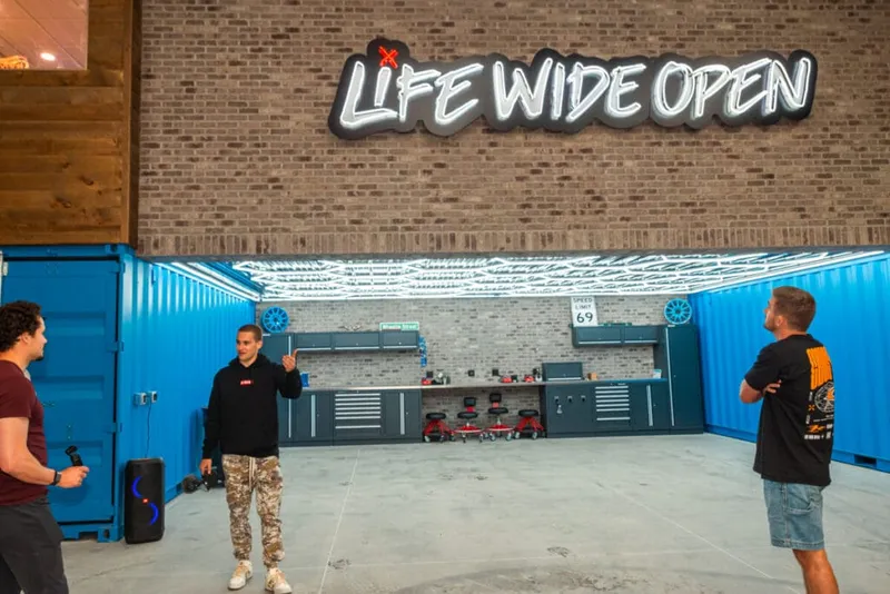 Three men stand inside a garage-style space beneath a lit 'Life Wide Open' sign on a brick wall