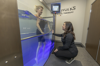 Woman walks in underwater treadmill tank while therapist crouches beside it