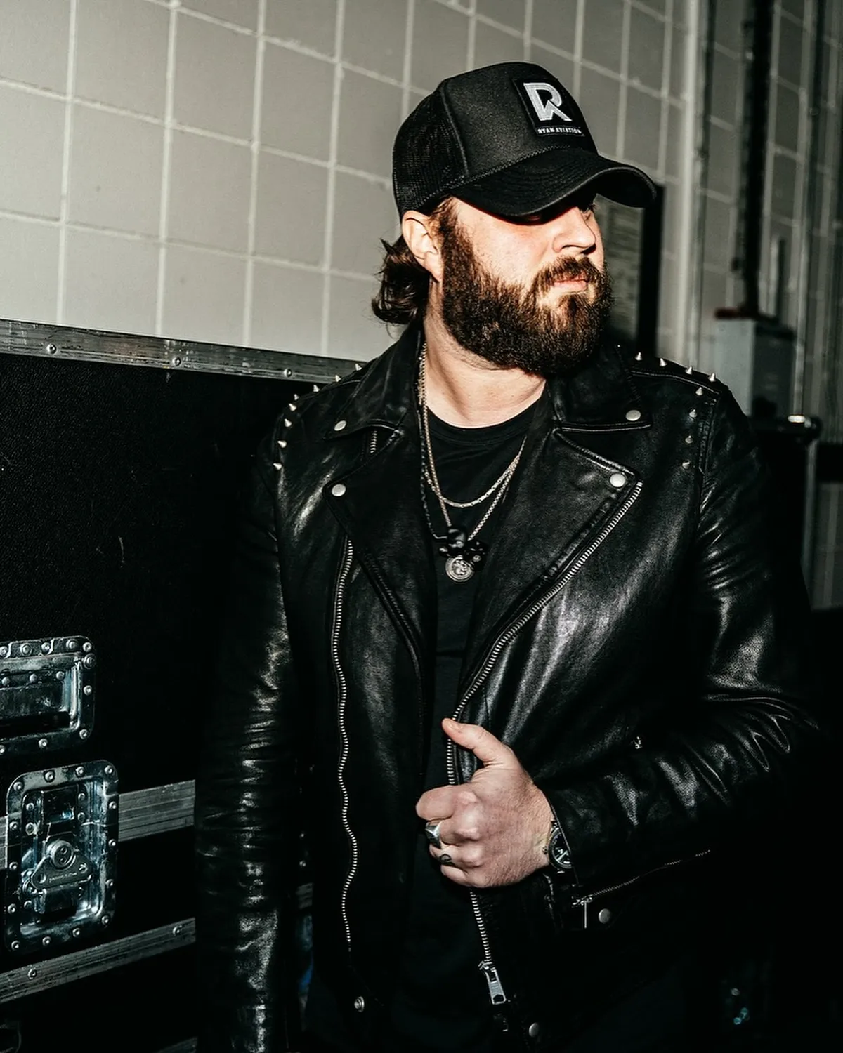 Bearded man in spiked leather jacket and black trucker cap standing backstage near road cases