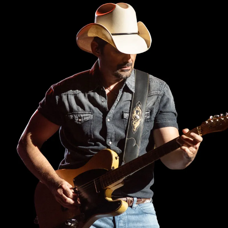 Man in white cowboy hat and denim shirt playing an electric guitar against a black background