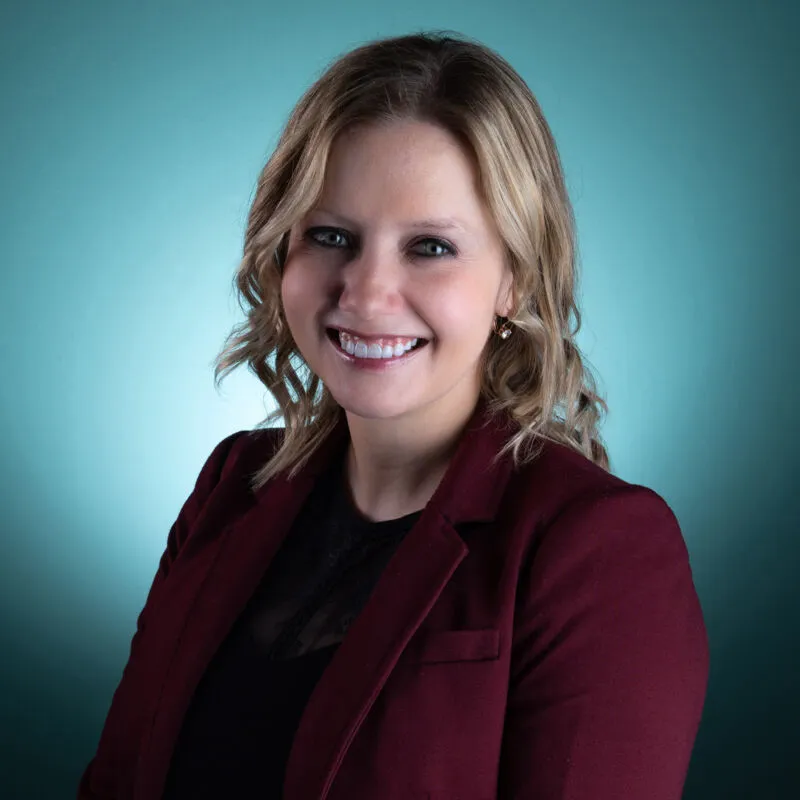 Smiling woman with blonde wavy hair wearing a dark red blazer against a teal background