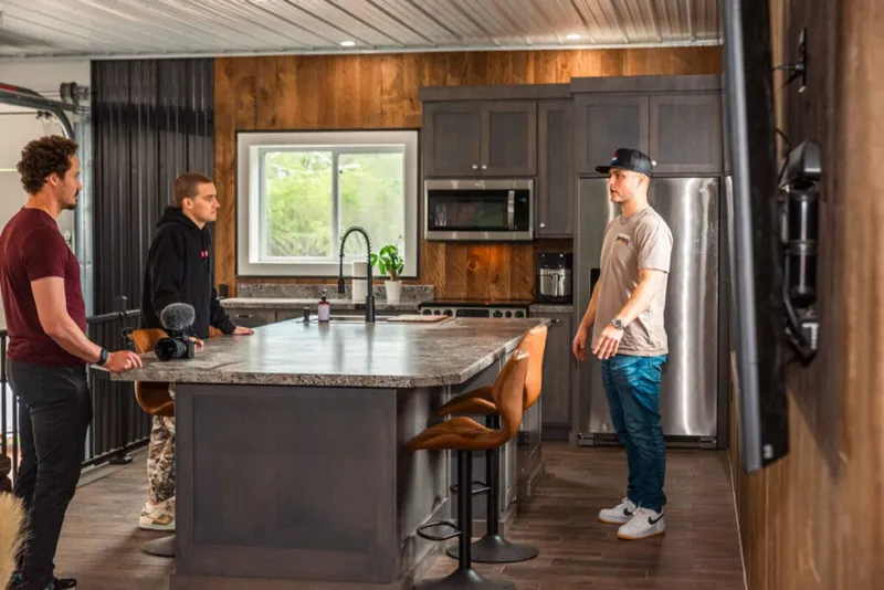 Three men gathered around a gray kitchen island during a video shoot, camera visible