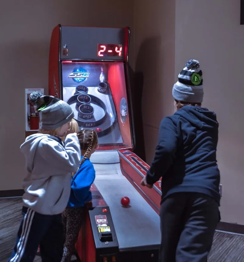 Two kids and an adult in winter hats playing a skee-ball arcade game
