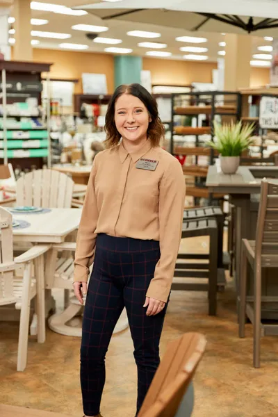 Smiling store employee in tan shirt with name badge stands among furniture displays