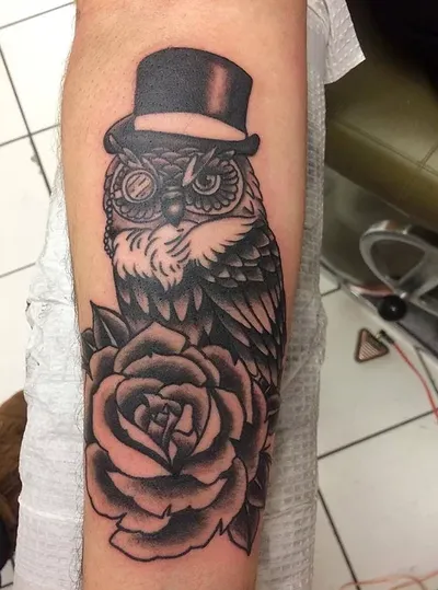 Black and grey forearm tattoo of an owl wearing a top hat above a rose