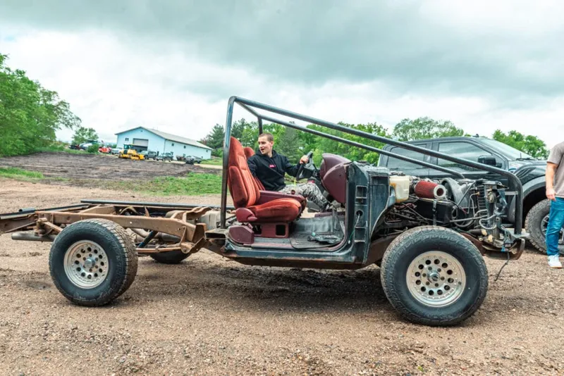 Young man grinning behind the wheel of a stripped bare-frame vehicle on a dirt lot