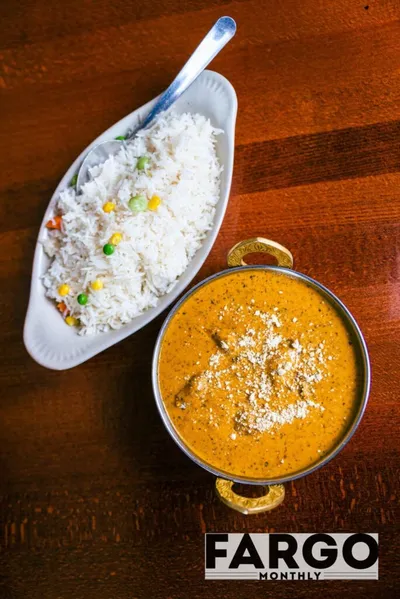 Overhead view of a creamy orange curry in a brass-handled bowl beside vegetable rice on a wooden table