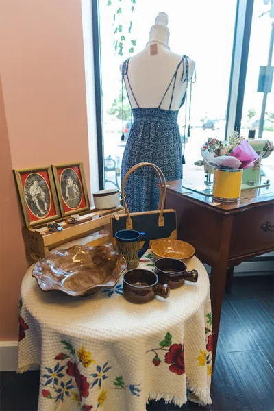 Vintage ceramics and handbag on a floral tablecloth beside a mannequin in a blue sundress in a boutique