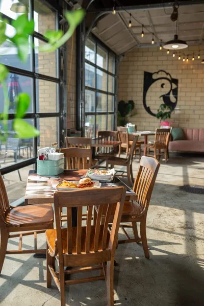 Sunny restaurant dining room with wood chairs, food-laden table, string lights, and a rooster logo on a brick wall