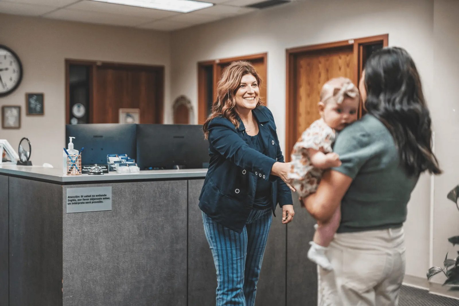 A smiling woman greets a mom holding her baby at a front desk