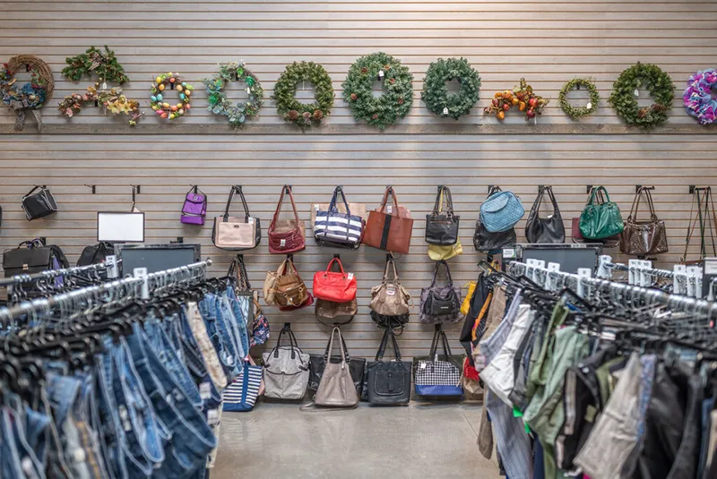 Thrift store wall displaying assorted handbags and holiday wreaths above two clothing racks