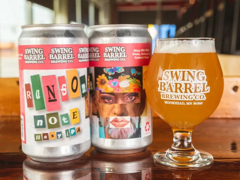 Two Swing Barrel Brewing Co. Ransom Note Hazy IPA cans beside a branded tulip glass of golden beer on a wooden bar