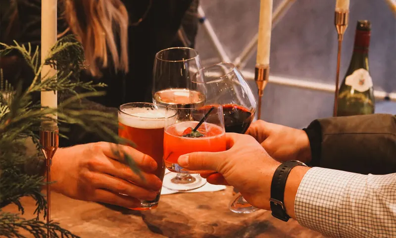 Three hands clinking beer, a cocktail, and red wine over a candlelit table with evergreen branches