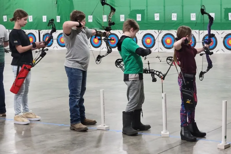 Four children aim compound bows at colorful target boards in an indoor archery range