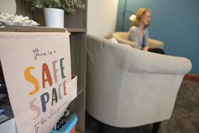 This is a Safe Space" sign in foreground with a person seated in a gray chair in a softly lit room