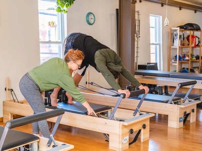 Pilates instructor guiding a student on a reformer machine in a bright wood-floored studio