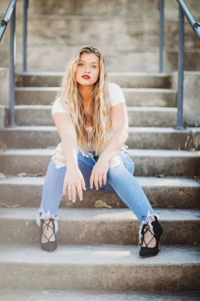 Young woman with long blonde hair sits on concrete steps in ripped jeans and black lace-up heels